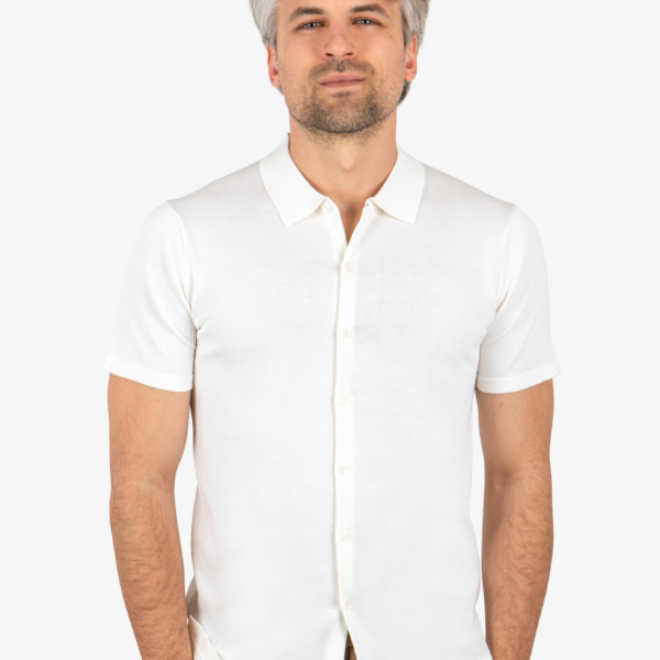 Man wearing white knit button shirt, standing with hands in pockets and facing forward.